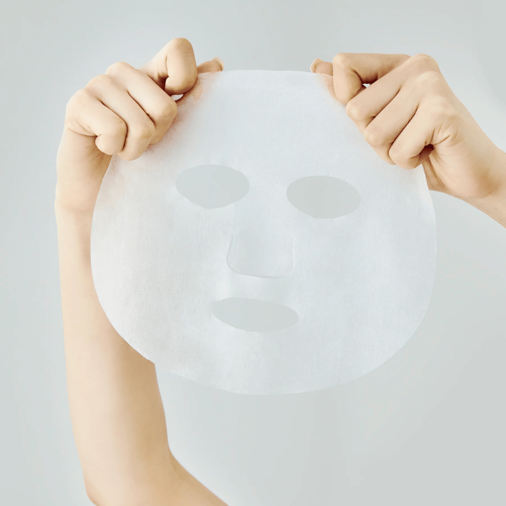 Två händer håller upp en vit arkmask mot en ljusgrå bakgrund, med synliga utskärningar för ögon, näsa och mun.