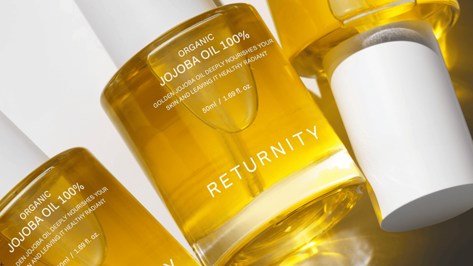 Närbild på flera flaskor med RETURNITY Organic Jojoba Oil 100% i transparent gul design. Bilden framhäver den gyllene jojobaoljan, stilren förpackning och känslan av närande och återfuktande hudvård.