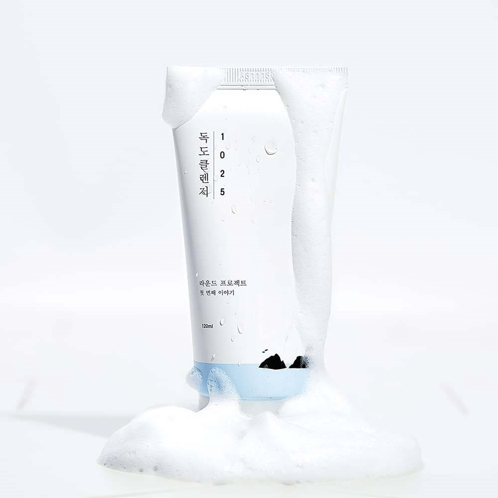 1025 Dokdo Cleanser in a white tube covered with lather, with water droplets and foam around the bottom, isolated against a light background.