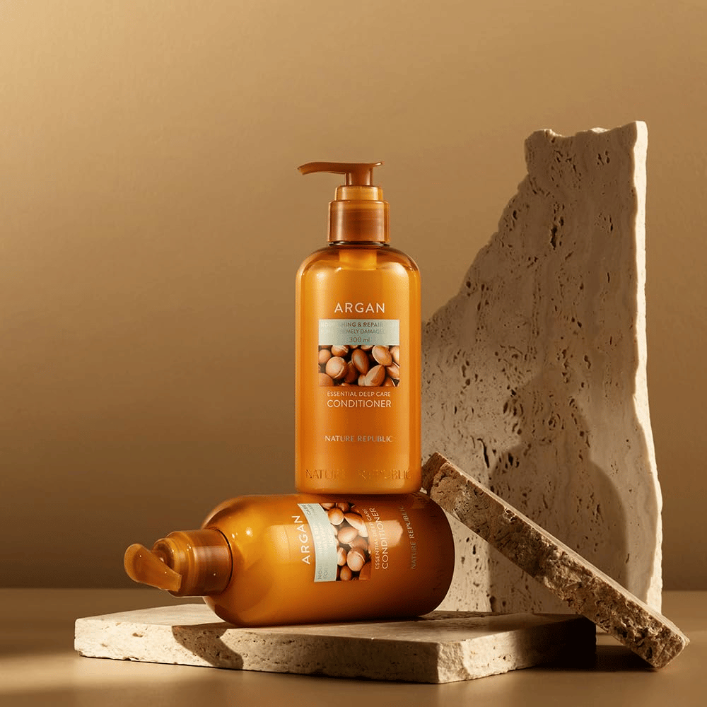 Two bottles of Nature Republic Argan Essential Moisture Repairing Conditioner on a warm-toned stone surface.