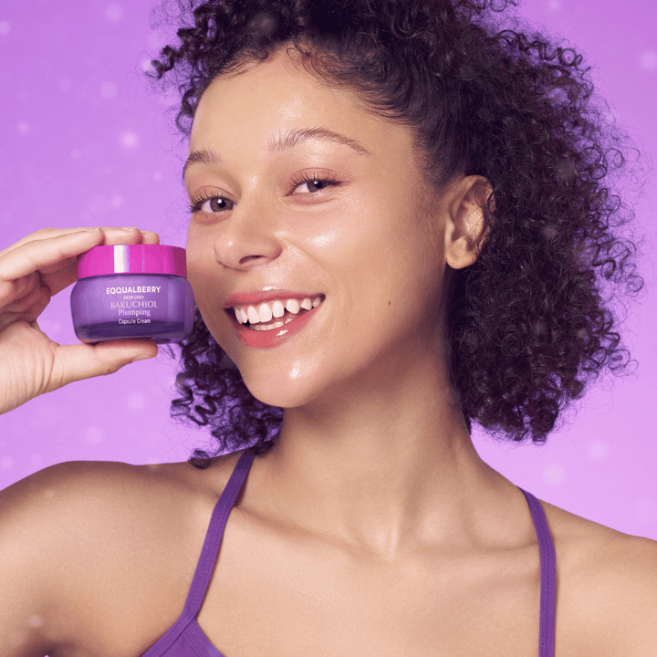 Kvinna med glödande hy håller EQQUALBERRY Bakuchiol Plumping Capsule Cream och ler framför en lila bakgrund.