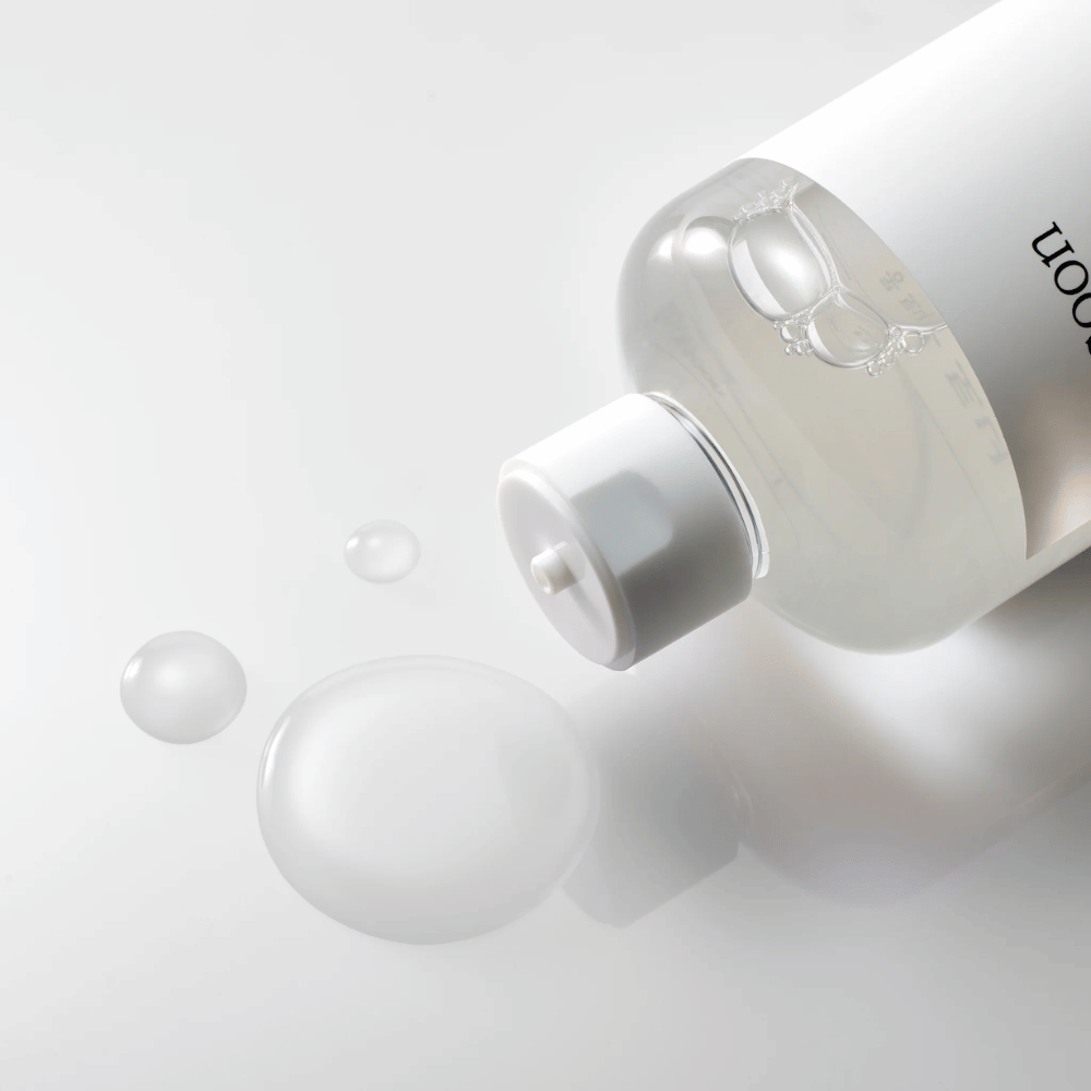 A transparent bottle lies on its side on a shiny surface with a light toner slowly flowing out, forming droplets and a larger pool on the surface. The bottle has a white label and a white pump mechanism. The lighting is soft, creating a clean and soothing atmosphere, while the toner’s fluid consistency and clarity are highlighted by how it spreads on the surface.