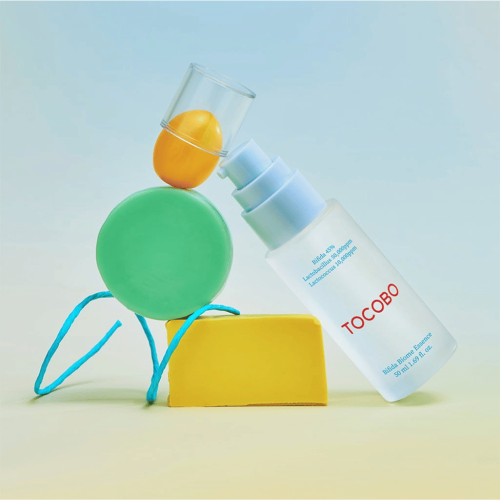 TOCOBO Bifida Biome Essence in a frosted pump bottle with a red logo, placed against a colorful background with green, yellow, and orange geometric shapes. Text on the bottle indicates Bifida 5%, Lactobacillus 3,000 ppm, and Lactococcus 1,000 ppm.