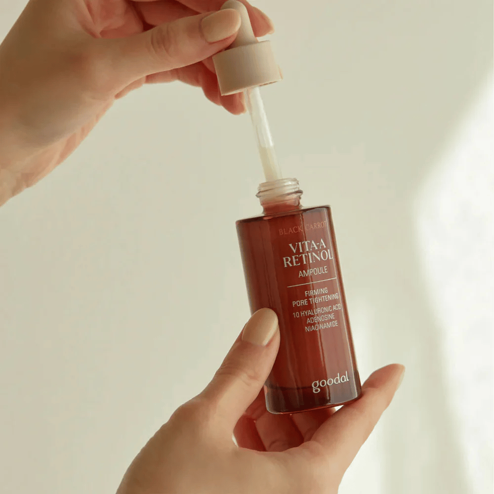 A hand holds a bottle with Goodal Black Carrot Vita-A Retinol Firming Ampoule. The bottle's label shows the product's name and features, including "Firming Pore Tightening" and ingredients like 10% hyaluronic acid, adenosine, and niacinamide. The dropper is beige in color.