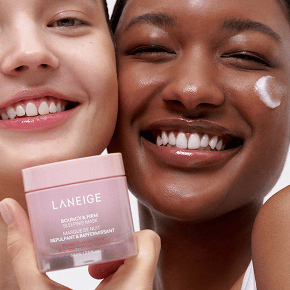 Two people smiling and holding Laneige Bouncy & Firm Sleeping Mask. One has applied the mask to their cheek, and the product is clearly visible in their hands. The mask is in a pink jar and marketed as "Plump, Hydrate, Glow."
