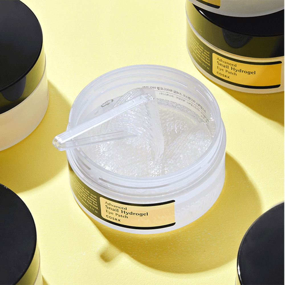 An open jar COSRX Advanced Snail Hydrogel Eye Patch with transparent eye patches and a small spatula, surrounded by more closed jars on a yellow background.