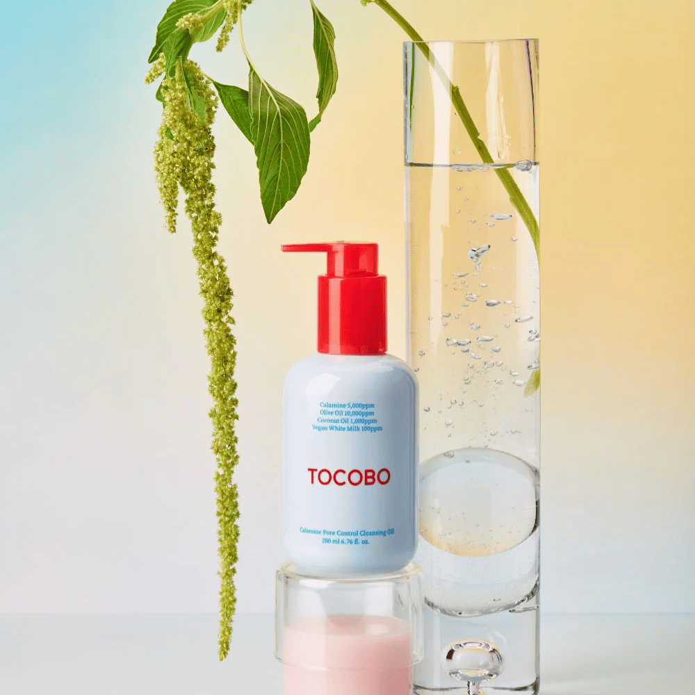 TOCOBO Calamine Pore Control Cleansing Oil with red pump stands next to a tall glass tube filled with water and a green plant, against a light pastel-colored background.
