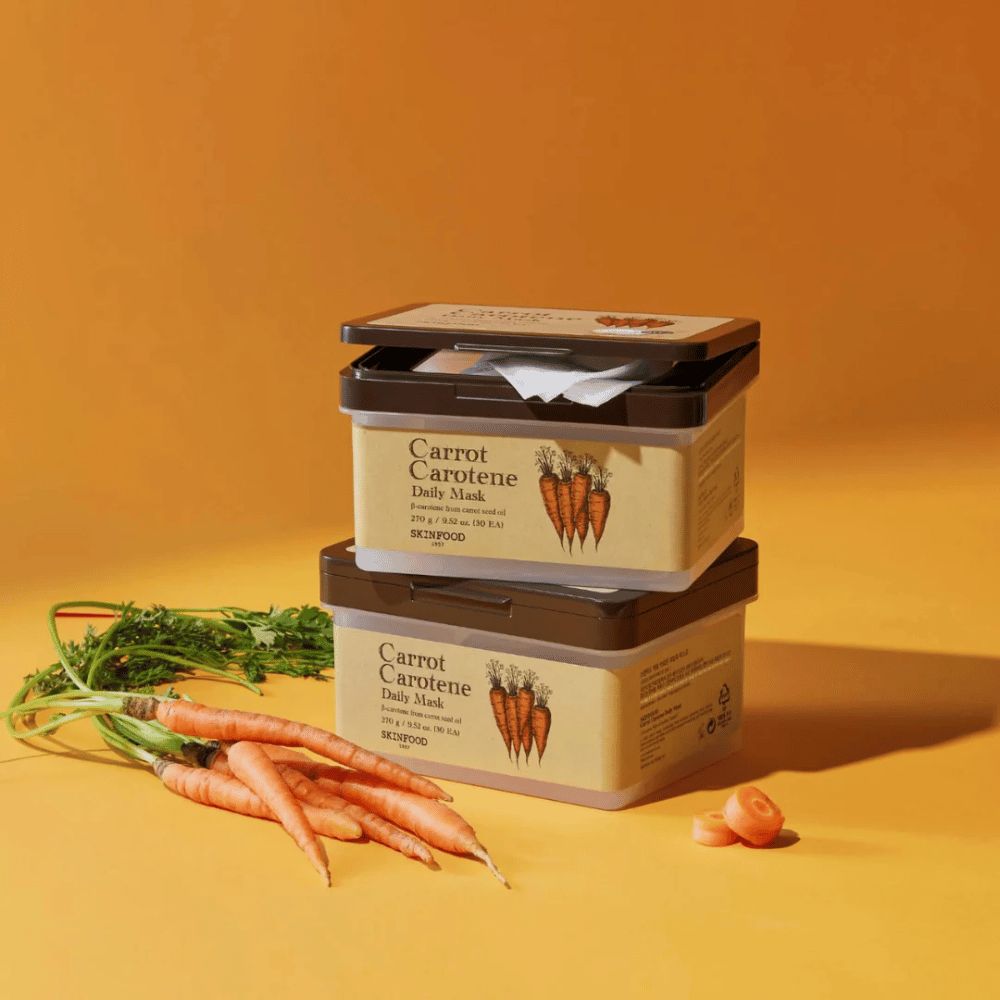 The image shows three stacked packages of "Carrot Carotene Daily Mask" from SKINFOOD, placed on an orange background along with fresh carrots. Each box contains face masks enriched with β-carotene from carrot seed oil, designed for daily use to nourish and care for the skin.