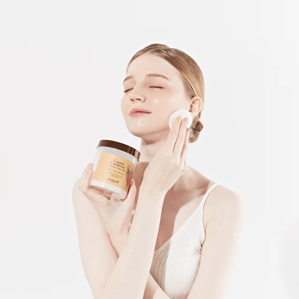 A woman with her hair up applies a skincare product to her face with a cotton pad. She holds a large jar labeled "Carrot Carotene Calming Water Pad". Her closed eyes and peaceful expression convey a sense of relaxation and enjoyment of the skincare routine.