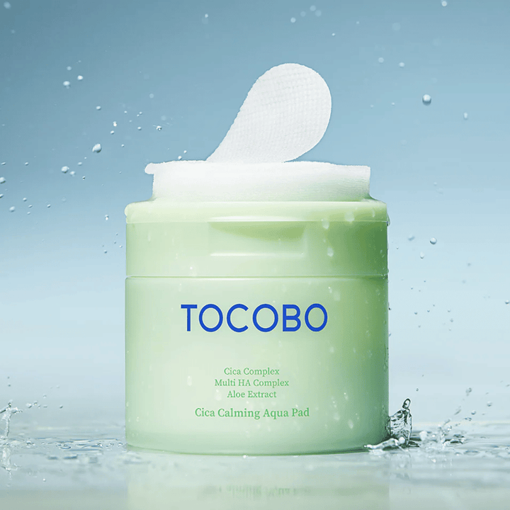 Open jar of TOCOBO Cica Calming Aqua Pad with a toner pad sticking up from the packaging. Water droplets around the product enhance the feeling of intense hydration and freshness.