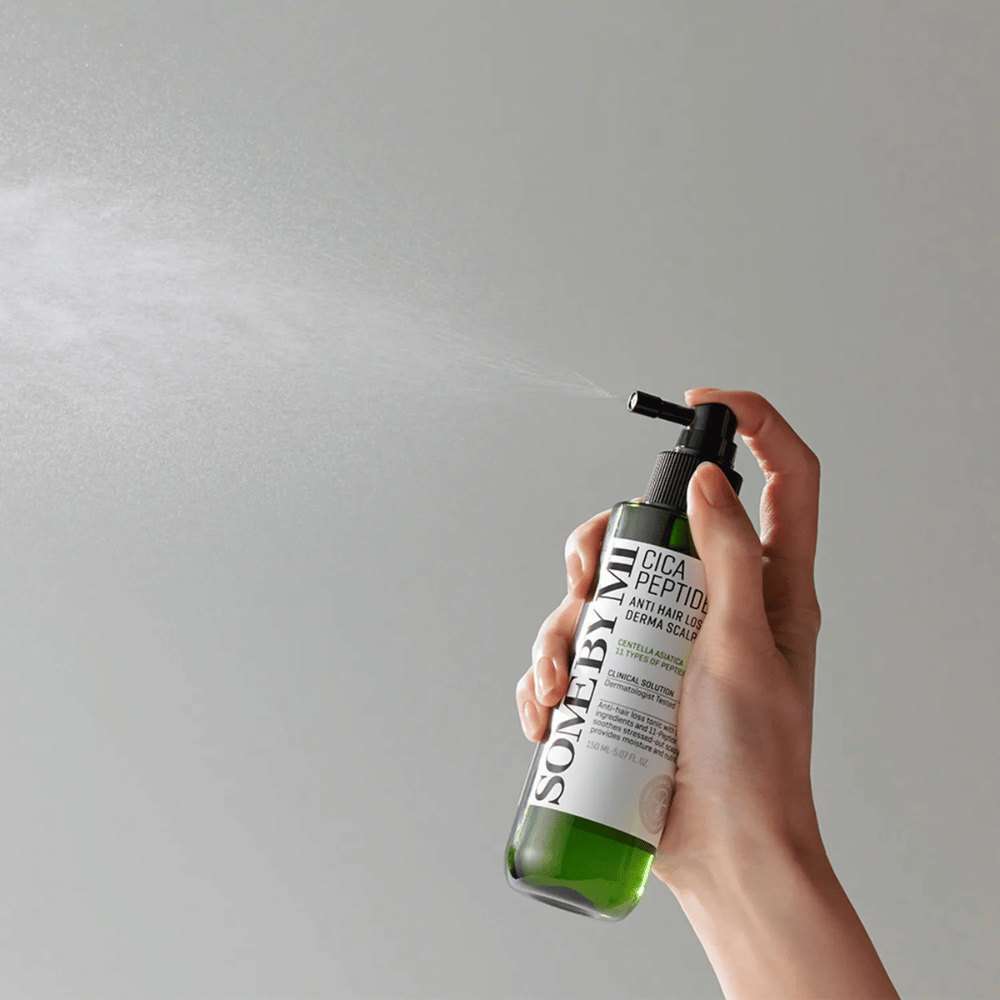 Hand spraying SOME BY MI Cica Peptide Anti Hair Loss Derma Scalp Tonic from a green bottle with black nozzle and white label against gray background.