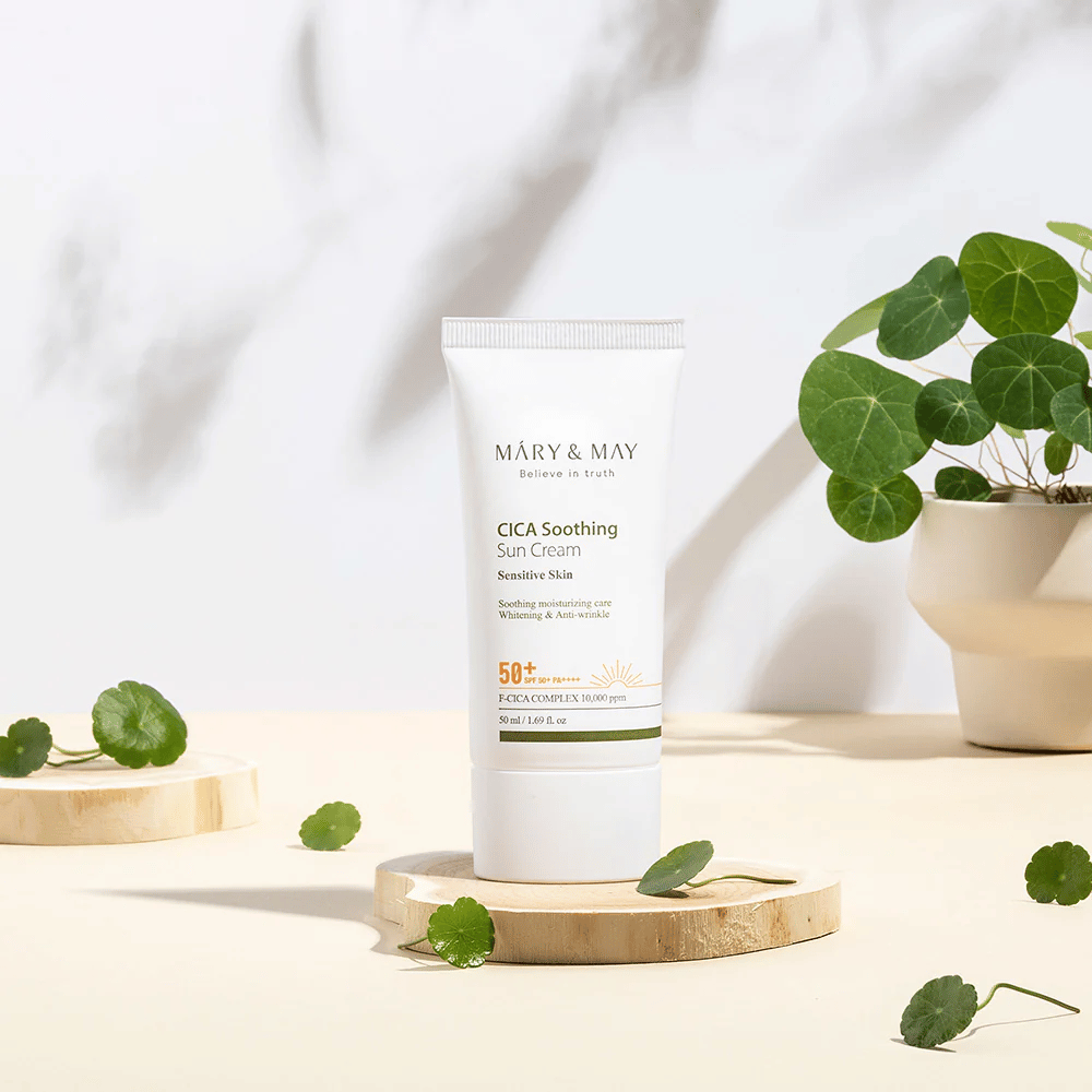 Mary&May CICA Soothing Sun Cream placed on a wooden podium with green centella leaves around it. The image emphasizes the skin-soothing ingredients and the natural inspiration behind the product.