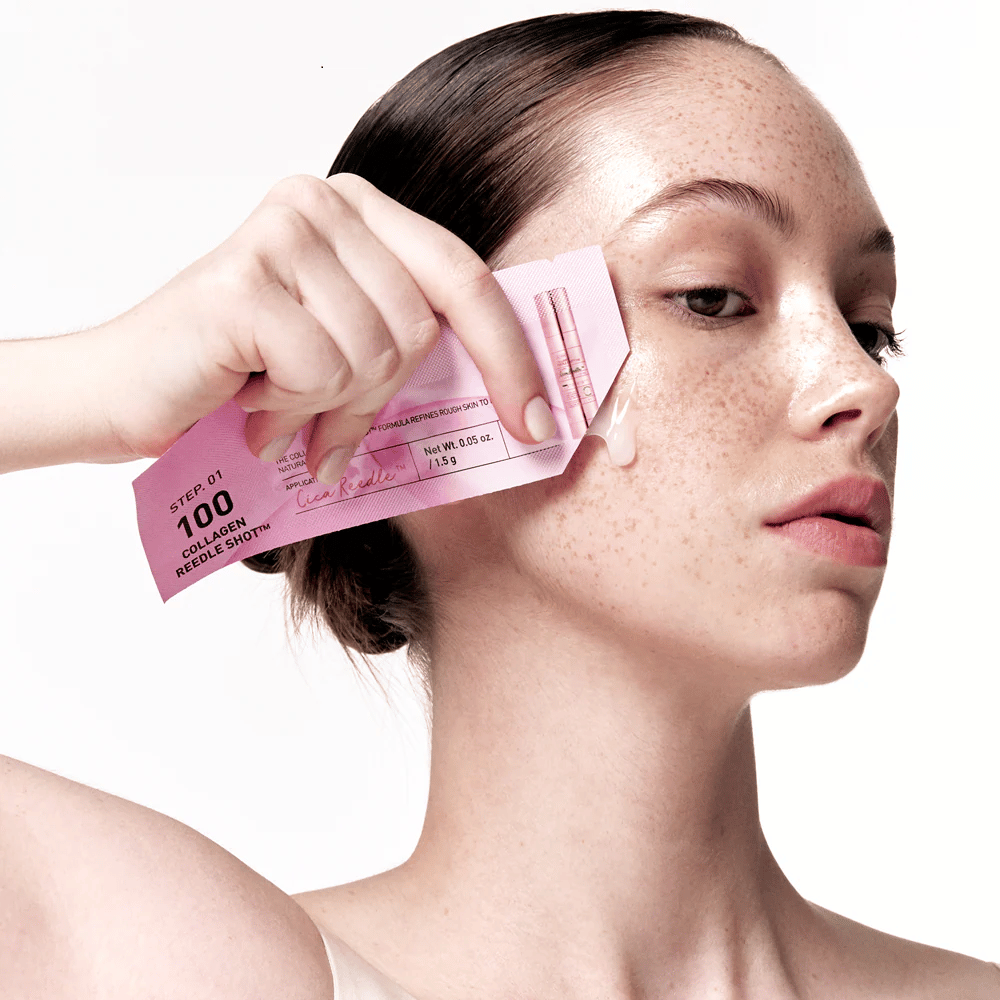 Woman applies step 1 of Collagen Reedle Shot 100 with a pink single-use packaging on the cheek. A clear gel is visible on the skin, marking the first step in the two-step skincare treatment.