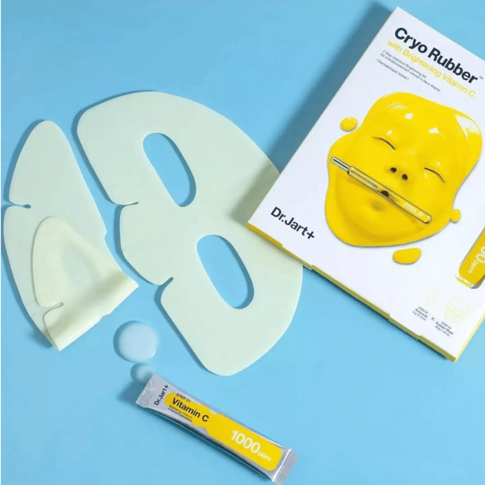 Image of Dr.Jart+'s 'Cryo Rubber™ with Brightening Vitamin C' beauty product. On a blue background, a white rubber face mask, a small tube labeled 'Vitamin C 1000ppm', and a box with the product's name and image of the yellow mask are seen.
