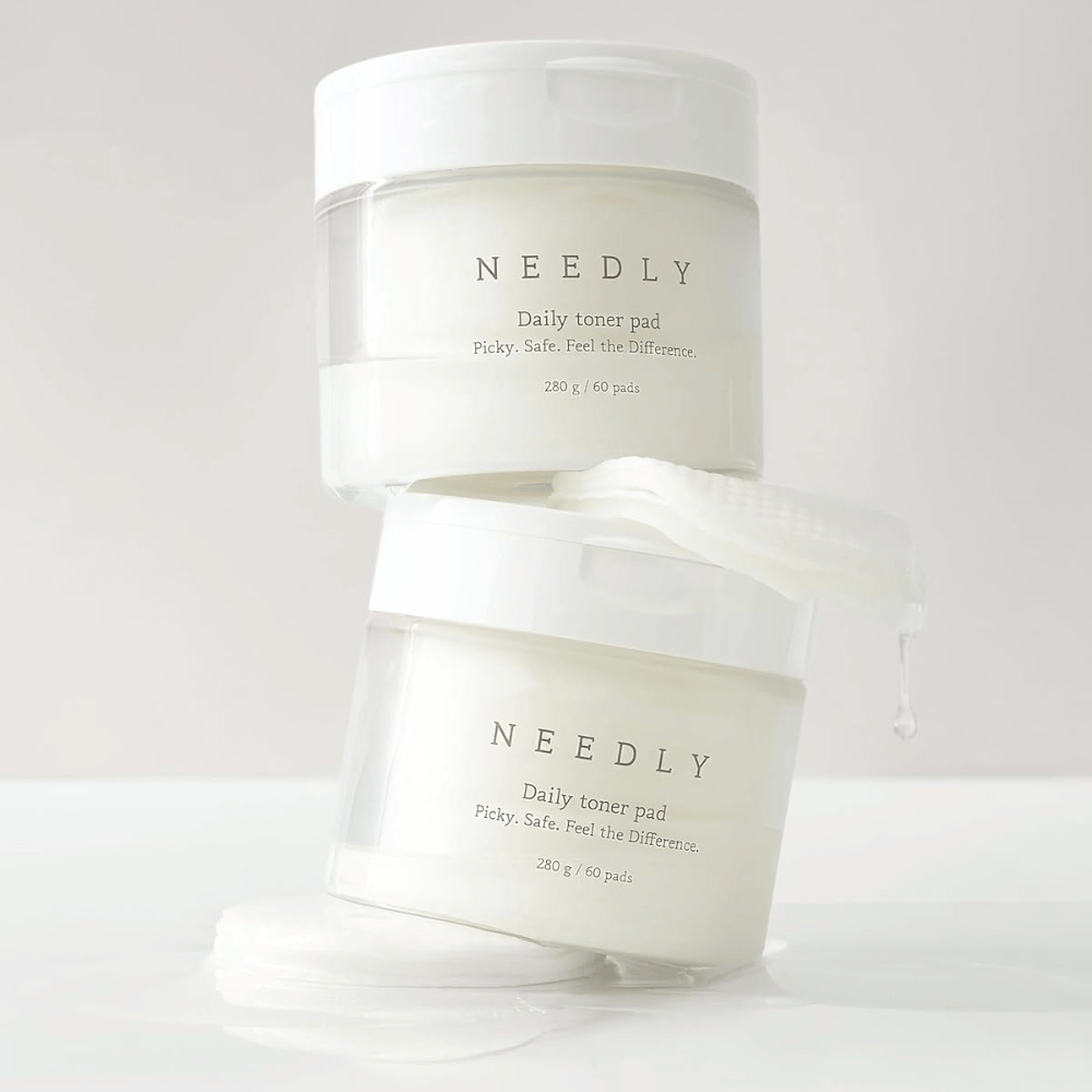 Two jars with Needly Daily Toner Pad stacked on top of each other. The jars are white with minimalist text and contain 60 toner pads each. A toner pad is partially outside the upper jar with liquid dripping off.