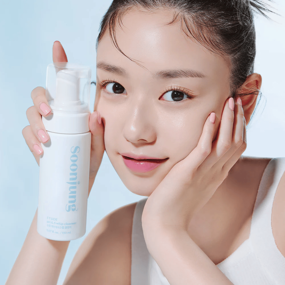 A young woman with clear skin holds a white pump bottle with the text "Soon Jung pH 6.5 Whip Cleanser" from ETUDE. She smiles at the camera with light makeup and a fresh appearance against a light blue background.