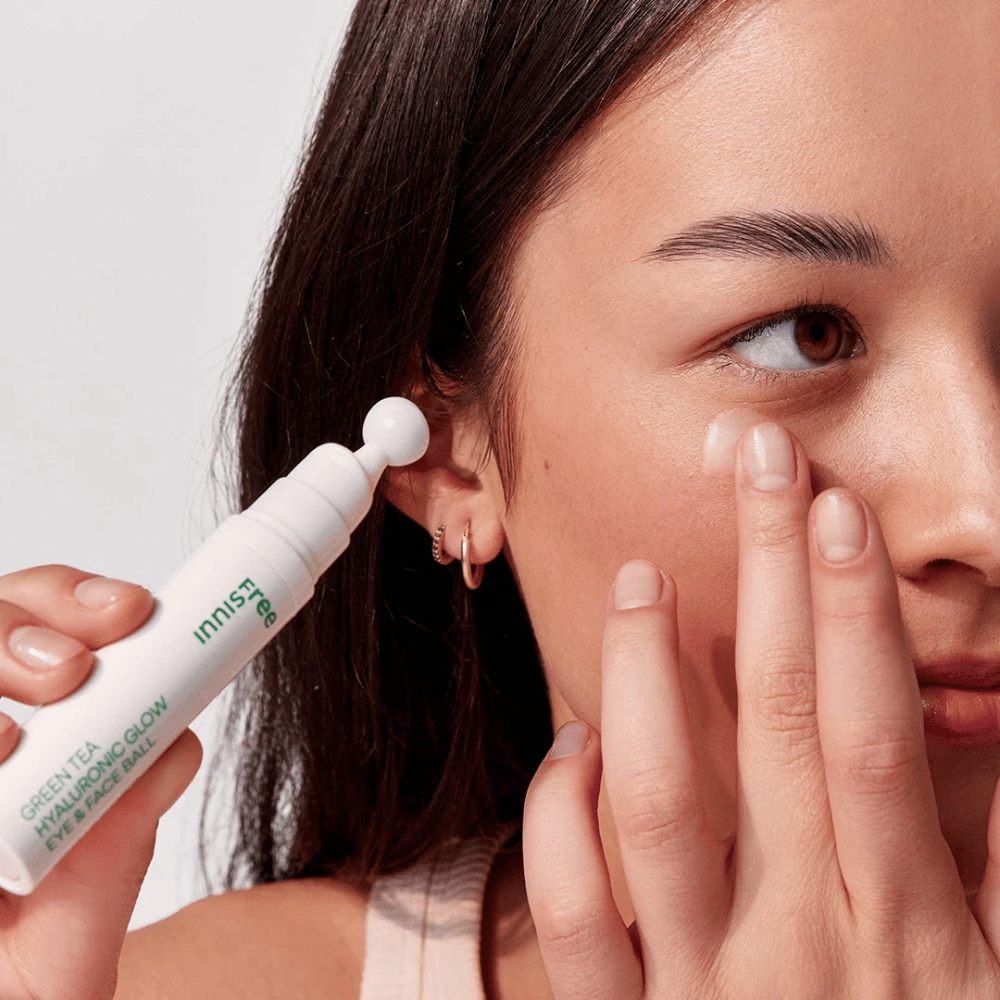 A person applies Innisfree Green Tea Hyaluronic Glow Eye & Face Ball on their skin under the eyes. The product has a white, round applicator and is in a white bottle with green text. The user's face is partially visible and focuses on the application of the product.