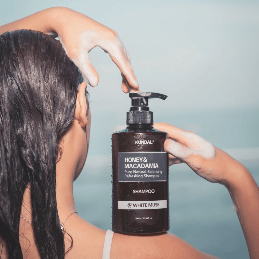 Woman with wet hair applies foam and holds KUNDAL Honey & Macadamia Nature Shampoo White Musk against a sea background.