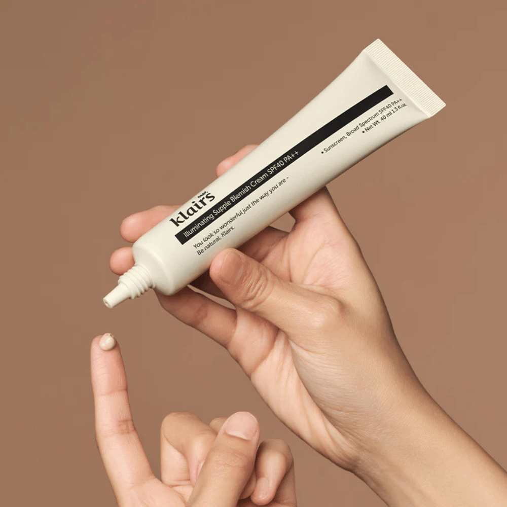 A hand holds a tube of Klairs Illuminating Supple Blemish Cream SPF40 PA++ and squeezes a small amount of cream onto a finger, against a brown background.