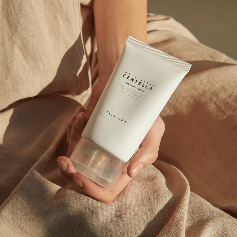 A person holds a white tube of Madagascar Centella Soothing Cream from SKIN1004. The tube has a transparent cap and text describing the product as soothing and made with pure Centella. The person is dressed in light-colored clothes.