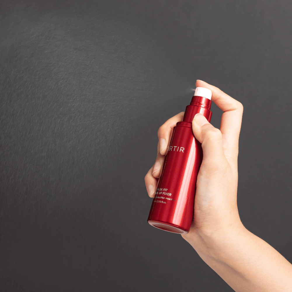 A hand holding a red bottle of brand tirtir Mask Fit Make Up Fixer and spraying a fine mist, ideal for setting makeup.