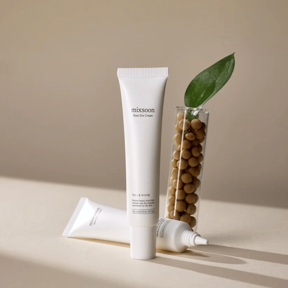 Two tubes of Mixsoon Bean Eye Cream placed next to a test tube filled with soybeans and a green plant leaf, on a beige surface with shadow play.