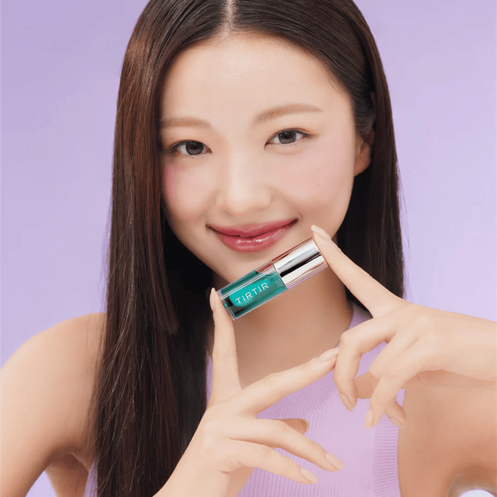 Woman with long, straight hair holding TIRTIR My Glow Lip Oil and showing off her glossy, pink lips against a purple background.