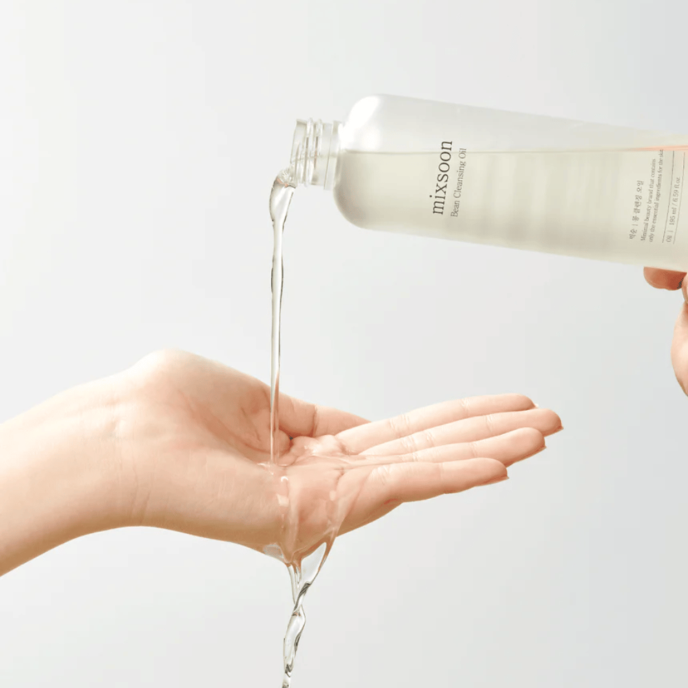 A clear cleansing oil is poured from a transparent bottle with a white label that reads "MixSoon" into an open palm. The oil forms a glossy stream between the bottle's opening and the hand, demonstrating the oil's liquid consistency. The hand is still and held so that it can collect the entire amount of cleansing oil against a plain light background.
