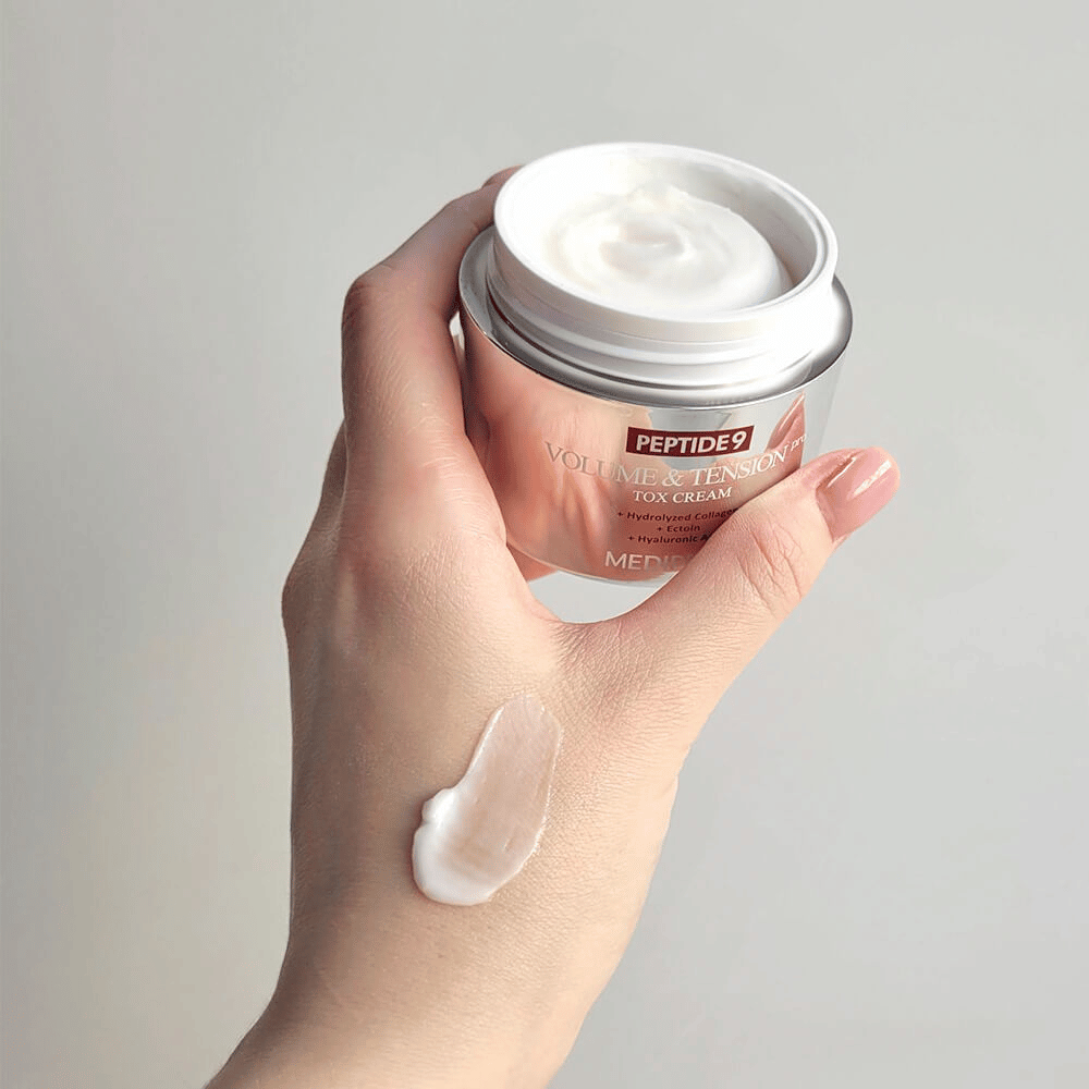 Hand holding MEDI-PEEL Peptide 9 Volume & Tension Tox Cream with a dab of cream on the back of the hand. The silky formula moisturizes and gives the skin firmness and elasticity.