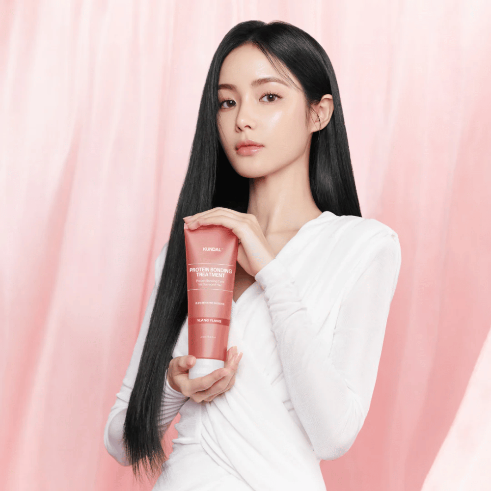 Woman with long, shiny hair holding KUNDAL Protein Bonding Treatment Violet Muguet against a pink background.