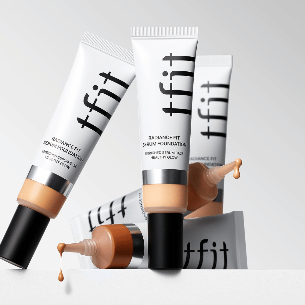 Several tubes of TFIT Radiance Fit Serum Foundation in different shades, one of which is dripping with foundation, against a light gray background.