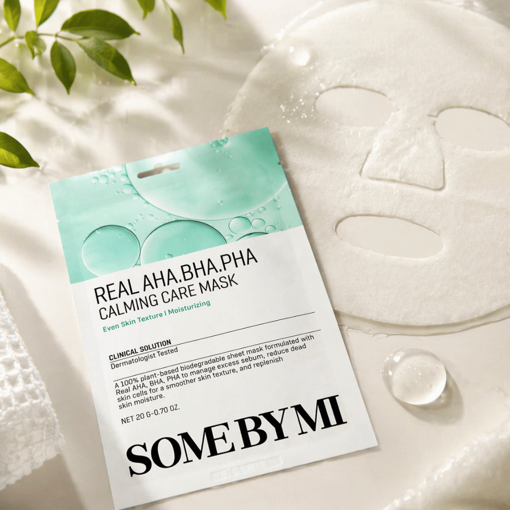 SOME BY MI Real AHA.BHA.PHA Calming Care Mask is shown together with the unfolded sheet mask. Around the product, moisture droplets and green leaves are visible, reinforcing the feeling of hydration, soothing care, and gentle exfoliation. The image highlights the mask's focus on a more even skin texture and improved moisture balance.