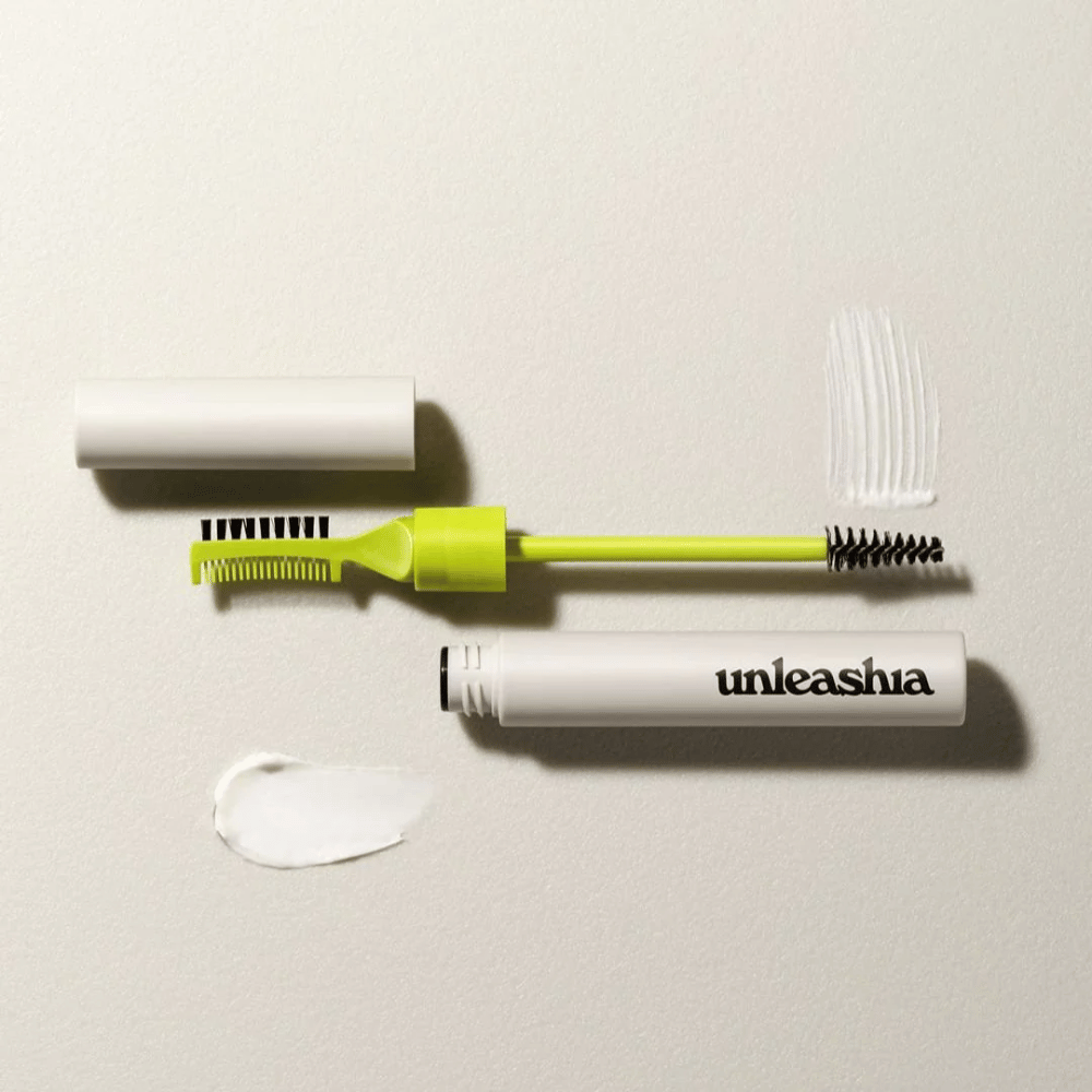 An eyebrow sculpting set with a green comb, black brush, and a white tube labeled "unleashia" on a light background with a clump of transparent product.