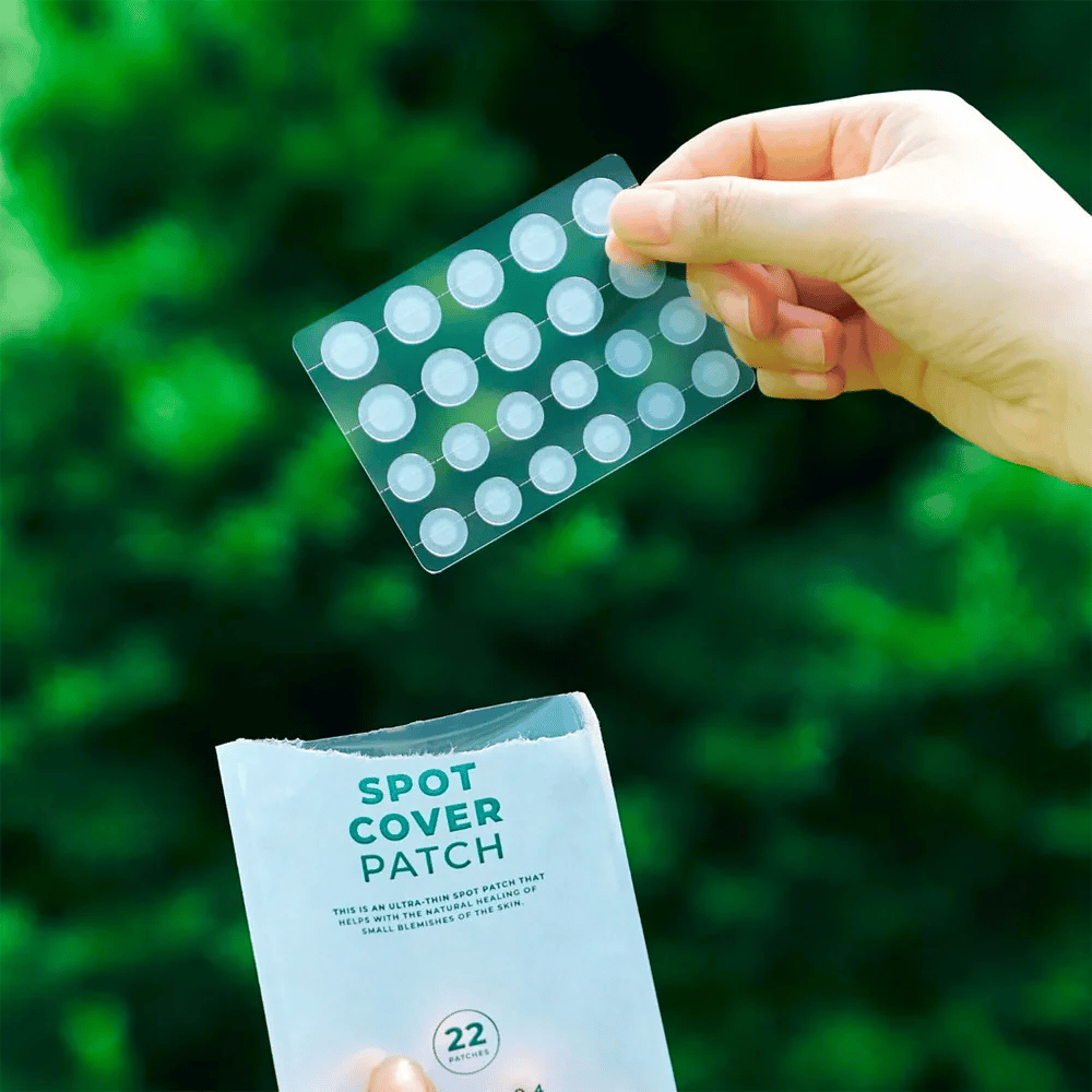 A hand holds up a sheet of Spot Cover Patches, which are transparent and rounded. In the background, a blurry green environment is visible. The packaging shows a part of the packaging with the text "SPOT COVER PATCH" and information that the product contains 22 patches that help heal small skin areas.