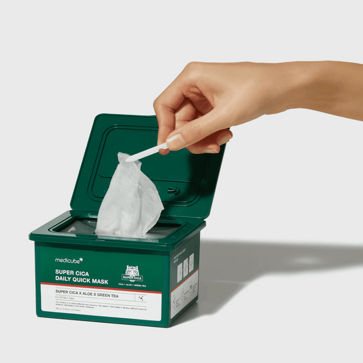 Grön behållare med öppet lock visar medicube Super Cica Daily Quick Mask. En hand drar upp en arkmask med medföljande pincett. Produkten innehåller Cica, aloe och grönt te, idealisk för snabb återfuktning och lugnande effekt.