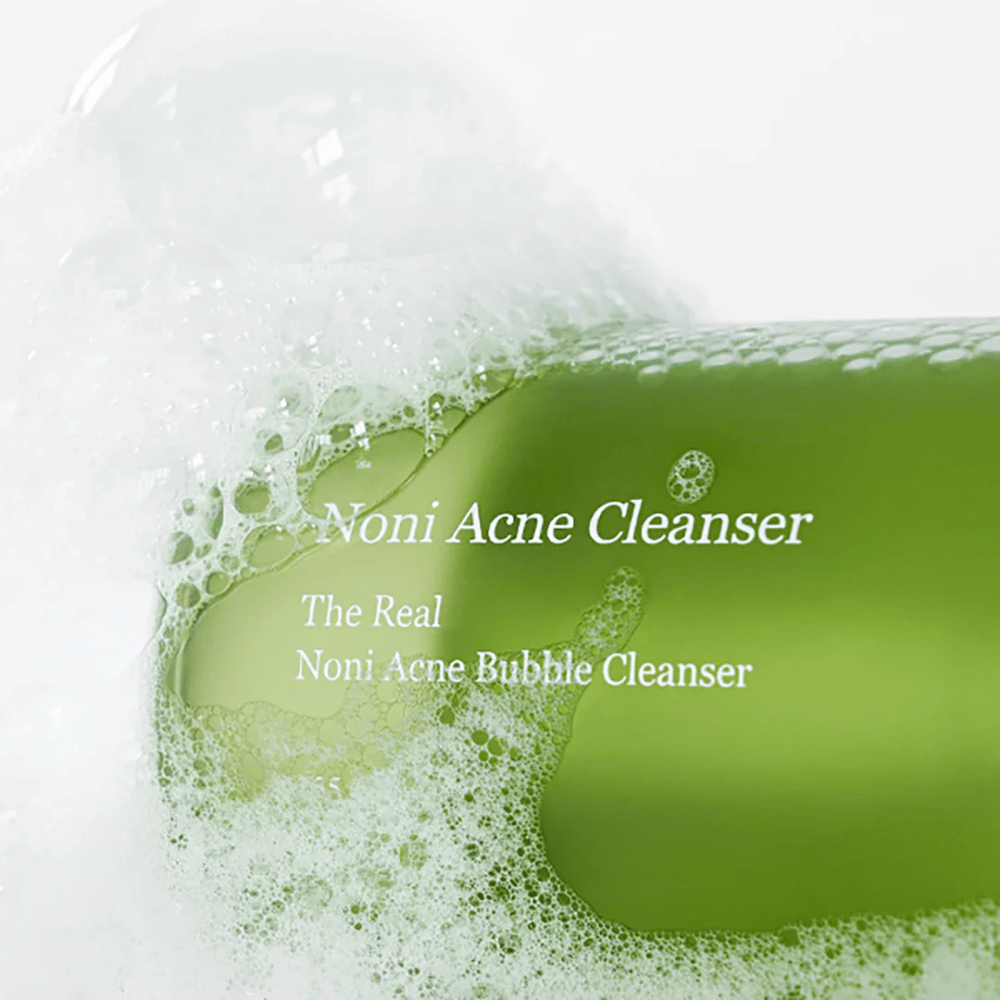 The bottle with The Real Noni Acne Bubble Cleanser is in focus and surrounded by plenty of foam. The fresh green color signals natural ingredients, perfect for cleaning and combating acne. An ideal product for clear and clean skin.