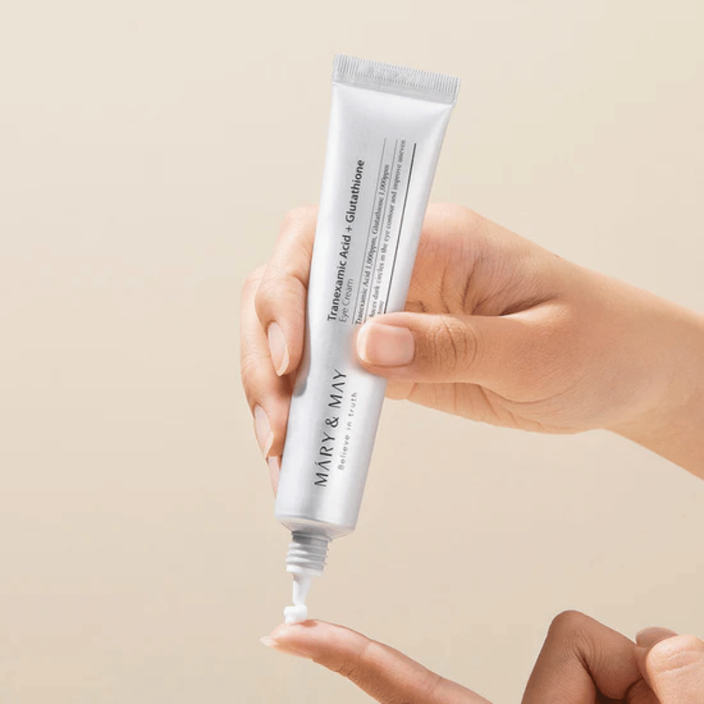 A hand holds a tube of MARY & MAY "Tranexamic Acid + Glutathione Eye Cream". The tube is white and the design is clean and simple, highlighting the product's soothing and brightening properties for the eye area. The product is applied gently, with a focus on precision and efficiency.