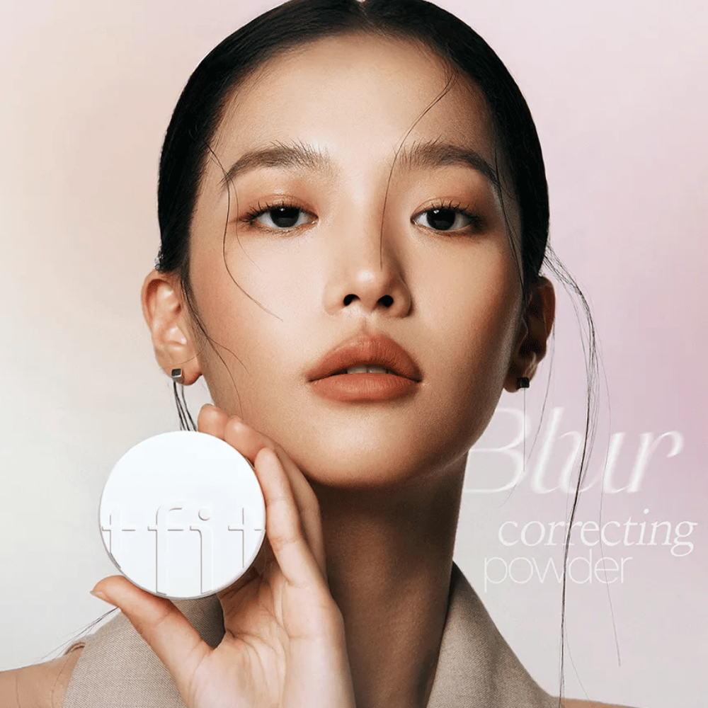 Woman with flawless makeup holding up a round white compact with TFIT logo, with the text "Blur Correcting Powder" in the background.