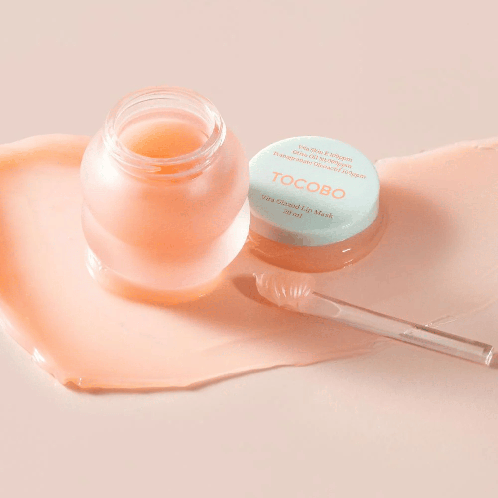 A jar of "Vita Glazed Lip Mask" from Tocobo is displayed. The jar is transparent with a light turquoise lid and contains a glossy, pink lip mask. A small spatula lies next to the jar, and part of the mask is spread on the surface. The product is designed to moisturize and care for the lips.
