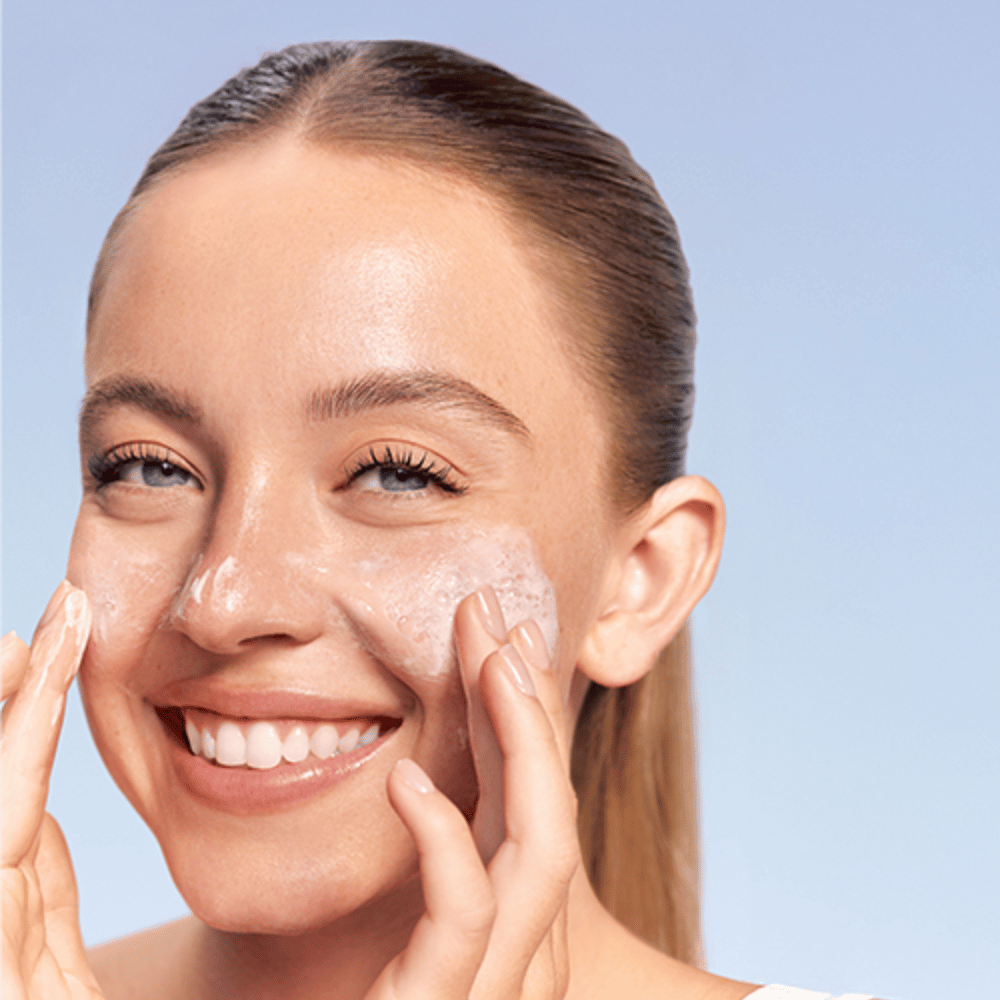 Smiling woman applies LANEIGE Water Bank Gentle Gel Cleanser in foaming form on her cheeks, against a blue background.