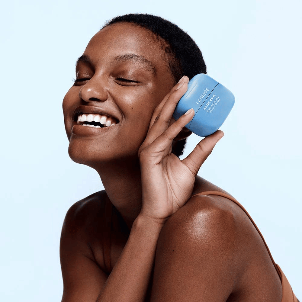 Smiling woman holds up a blue jar of LANEIGE Water Bank Blue Hyaluronic Cream against her cheek, with a light blue background.
