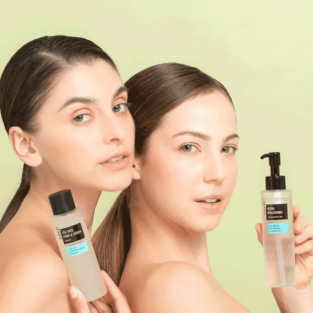 Two women with bare shoulders pose with skincare products; one holds a bottle labeled "TEA TREE PORE & SEBUM", and the other a pump bottle labeled "ULTRA HYALURONIC CLEANSING OIL". They stand in front of a light green background.